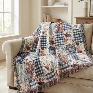 May include: A patchwork throw blanket on a beige sofa. The blanket has floral and gingham patterns in blue, cream, and burgundy. The edges are finished with a fringe. The sofa is in a living room.