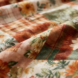 May include: Close-up of a patchwork fabric with floral and plaid patterns. The fabric features a mix of warm colors, including orange, brown, green, and cream. The design includes detailed floral prints and plaid squares, creating a cozy, textured appearance.