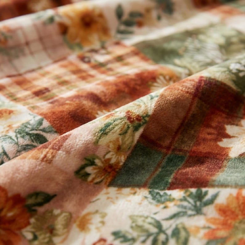 May include: Close-up of a patchwork fabric with floral and plaid patterns. The fabric features a mix of warm colors, including orange, brown, green, and cream. The design includes detailed floral prints and plaid squares, creating a cozy, textured appearance.