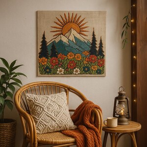 May include: Embroidered wall art featuring a mountain scene with a rising sun, evergreen trees, and colorful flowers. The artwork is displayed above a wicker chair with a cream-colored pillow and an orange blanket. A lantern and mug sit on a side table.