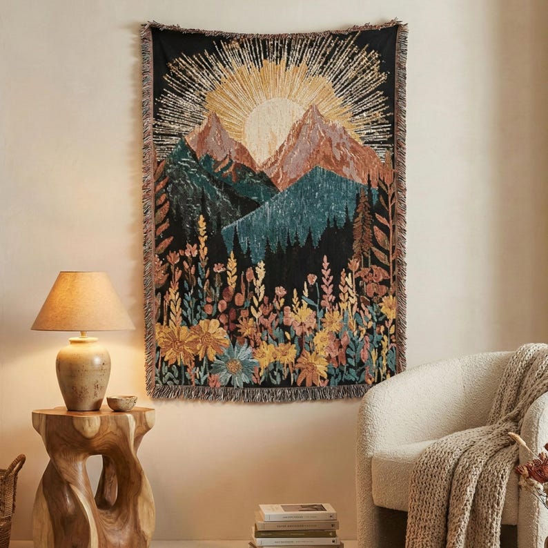 May include: A black tapestry with a mountain and sun design. The tapestry features a sunrise over mountains, with a forest and wildflowers in the foreground. The tapestry has a fringed edge and is hanging on a wall.