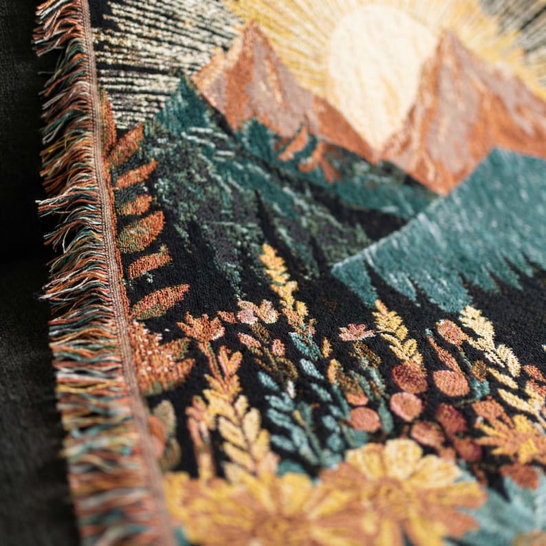 May include: A woven tapestry featuring a mountain landscape with a rising sun. The design incorporates shades of brown, green, and yellow, with a fringe border. The tapestry depicts a scenic view of mountains, trees, and wildflowers.