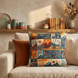 Puede incluir: Cojín decorativo cuadrado con un diseño de patchwork con escenas de montañas y flores en tonos naranja, azul y verde. Un cojín naranja liso está al lado en un sofá beige. Decoración del hogar.