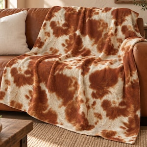 May include: A soft throw blanket featuring a realistic cowhide pattern with irregular patches of warm reddish-brown and creamy off-white colors. The fabric has a textured appearance that mimics the look of animal fur draped over a surface.