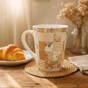 May include: A white ceramic coffee mug with a patchwork design in shades of beige, tan, and yellow, featuring floral and striped patterns. The mug sits on a woven coaster, next to a croissant on a white plate.