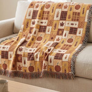Puede incluir: Una manta tejida con un diseño de patchwork de motivos otoñales que incluyen calabazas, varias formas de hojas y patrones de cuadros en tonos naranja, amarillo, marrón y crema, terminada con un borde de flecos en la parte inferior.