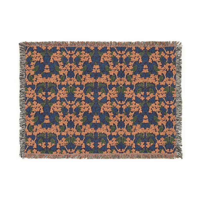 May include: A rectangular woven throw blanket featuring a repeating floral pattern with peach-colored blossoms and dark green leaves set against a deep navy blue background. The edges of the blanket are finished with a decorative fringed border.