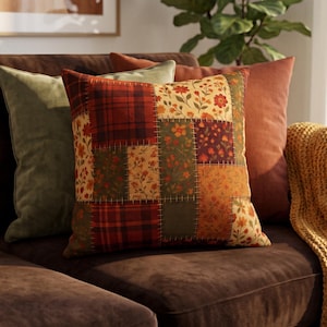 Puede incluir: Un cojín cuadrado con un diseño de patchwork compuesto por varios cuadrados de tela. El patrón incluye cuadros rojos y verdes, tela color crema con estampados florales rojos y naranjas, y secciones de color verde oscuro, unidas por costuras visibles.