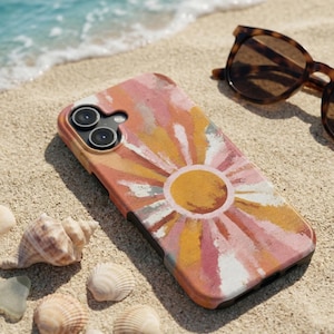 May include: A phone case with a colorful sunburst design in shades of pink, yellow, and orange. The case is lying on a sandy beach next to sunglasses and seashells. The case has a black border and a camera cutout.