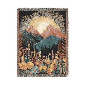 May include: A woven throw blanket featuring a mountain landscape with a rising sun. The design includes mountains in shades of brown, green, and blue, with a black background. The bottom of the blanket has a floral design in yellow, pink, and blue.