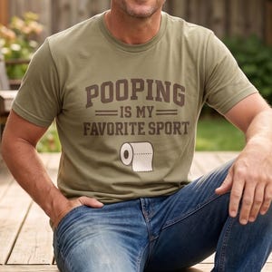 May include: An olive green short-sleeved t-shirt featuring the brown text POOPING IS MY FAVORITE SPORT in a bold, blocky font, accompanied by a graphic illustration of a white roll of toilet paper centered below the text on the chest.
