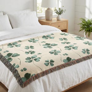 Peut inclure: Une couverture crème avec un motif répété de trèfles à quatre feuilles verts. La couverture a une bordure à franges et est présentée sur un lit blanc. Les trèfles sont de différentes tailles et sont reliés par de fines tiges vert foncé.