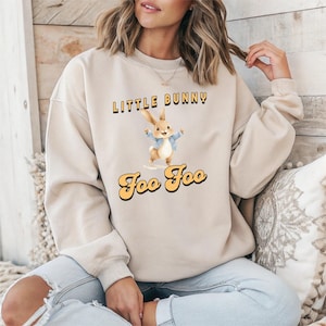 May include: Beige sweatshirt with the text "Little Bunny Foo Foo" in black and yellow lettering. The graphic features a cartoon bunny wearing a blue jacket. The sweatshirt is a neutral color.