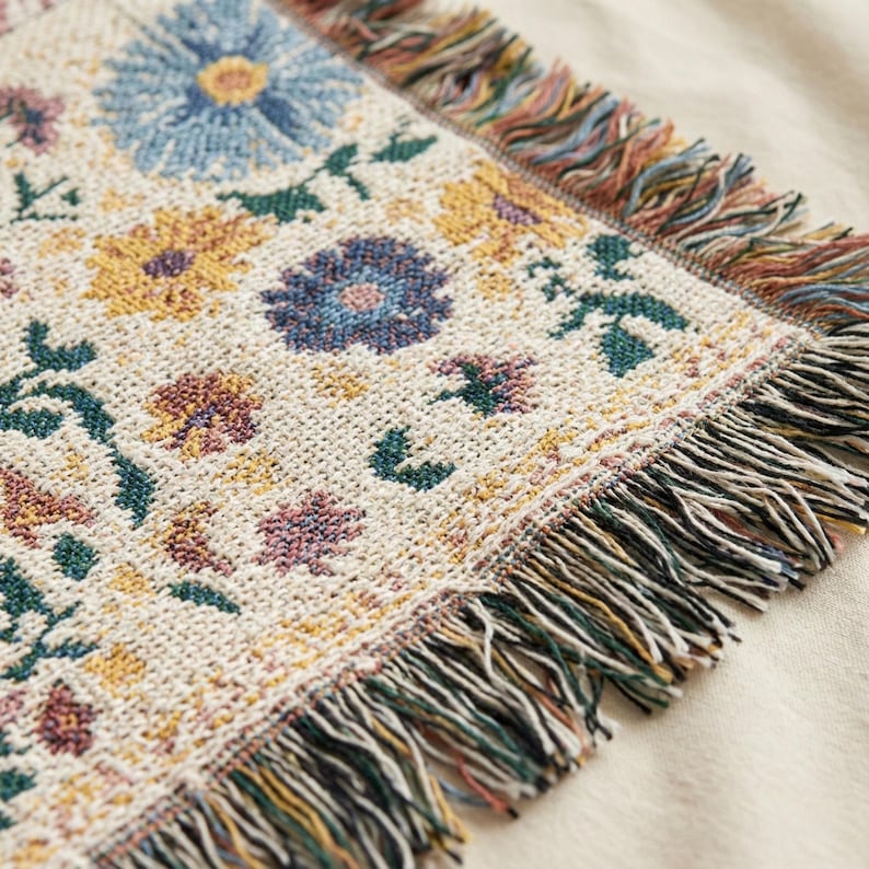 May include: Close-up of a woven floral throw blanket. The blanket features a cream-colored background with a repeating pattern of colorful flowers in shades of blue, yellow, and purple. The edges are finished with a fringe in coordinating colors.