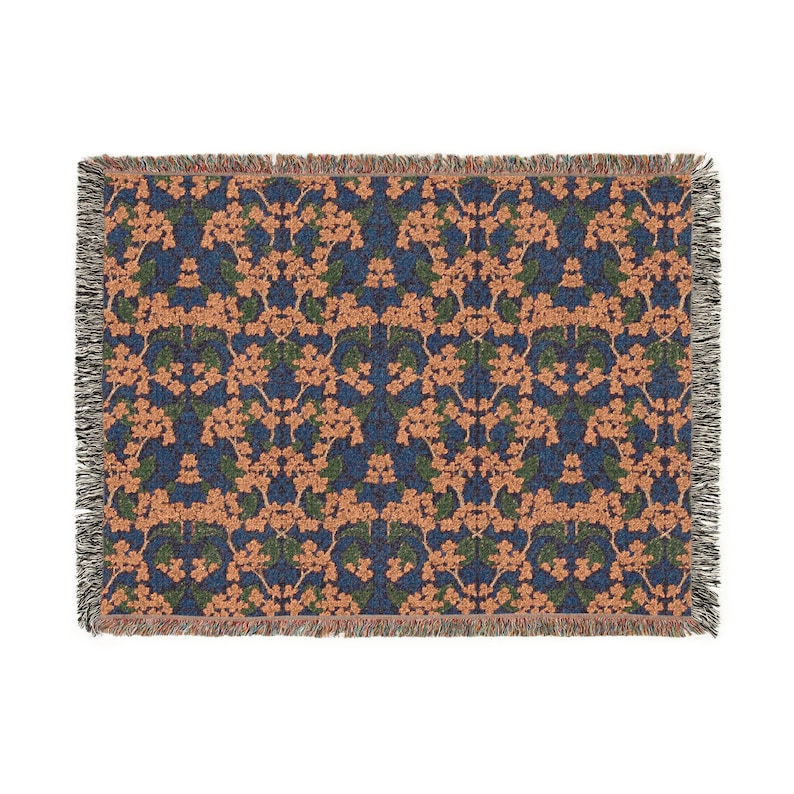 May include: A rectangular woven throw blanket featuring a repeating floral pattern with peach-colored blossoms and dark green leaves set against a deep navy blue background. The edges of the blanket are finished with a decorative fringed border.