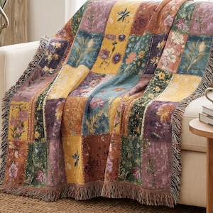 Puede incluir: Una manta tejida con un diseño de patchwork de paneles rectangulares con diversas ilustraciones florales y botánicas en tonos amarillo mostaza, verde oscuro, naranja óxido y morado apagado, con un borde acabado en flecos.