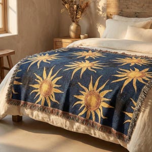 May include: A rectangular woven throw blanket featuring a repeating pattern of yellow and orange suns against a deep navy blue background. The blanket has a textured appearance with frayed edges along the sides and is draped over a bed.