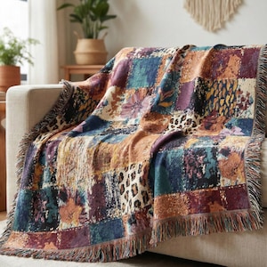 May include: A colorful patchwork throw blanket draped over a beige sofa. The blanket features squares of various patterns, including floral, leopard print, and abstract designs in shades of blue, purple, orange, and brown. The edges are finished with a fringe.