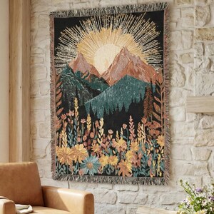 May include: A woven tapestry featuring a mountain landscape with a rising sun and a field of wildflowers. The tapestry has a black background with shades of green, brown, yellow, and blue. The tapestry is rectangular with a fringed edge.