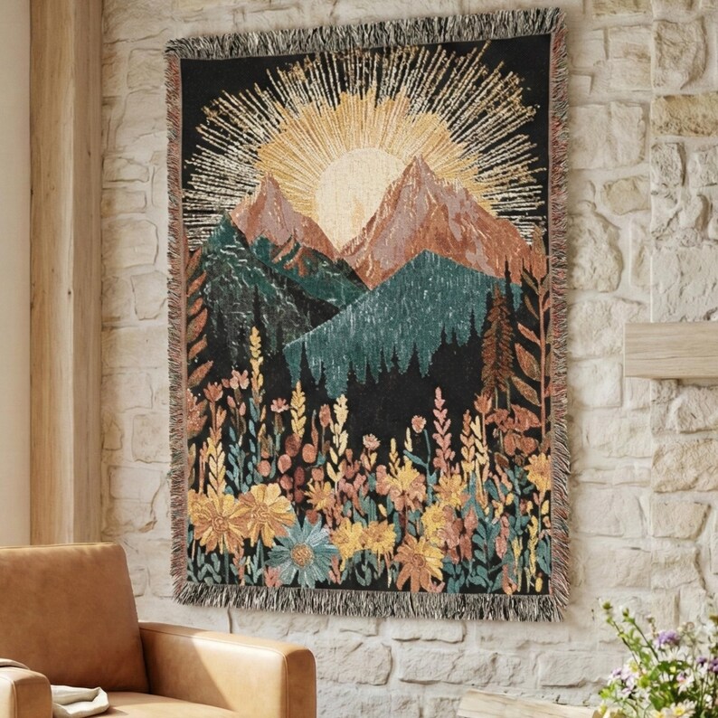 May include: A woven tapestry featuring a mountain landscape with a rising sun and a field of wildflowers. The tapestry has a black background with shades of green, brown, yellow, and blue. The tapestry is rectangular with a fringed edge.