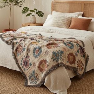 K&ouml;nnte beinhalten: Eine beige Tagesdecke mit einem Blumenmuster in Rot-, Blau-, Gelb- und Lilat&ouml;nen ist &uuml;ber ein Bett drapiert. Die Decke hat einen Fransenrand und liegt auf wei&szlig;er Bettw&auml;sche. Das Bett hat ein geflochtenes Kopfteil.