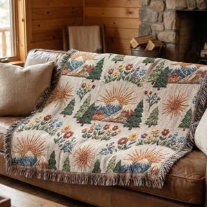 May include: A beige throw blanket with a repeating pattern of mountains, trees, and suns. The mountains are blue and the trees are green. The blanket has a fringed edge and is draped over a brown leather sofa.