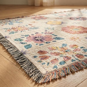 K&ouml;nnte beinhalten: Ein rechteckiger gewebter Teppich mit einem Blumenmuster in Pastellfarben. Der Teppich hat einen cremefarbenen Hintergrund mit Quadraten aus rosa, lila, gelben und blauen Blumen. Der Teppich hat einen Fransenrand.