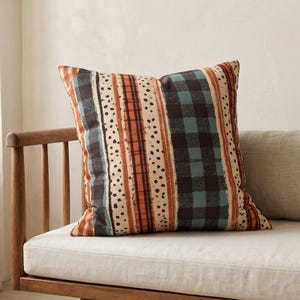 May include: A square throw pillow featuring a vertical pattern of alternating stripes. The design includes bands of black and teal buffalo check, solid orange lines, and cream-colored sections decorated with small, scattered black dots.