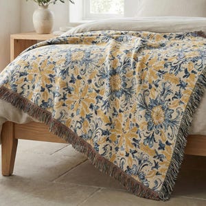 May include: A woven throw blanket with a floral pattern in yellow and blue on a cream background. The blanket has a fringed edge and is draped over a bed. The throw is made of a soft material.