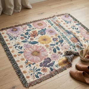 May include: A woven floral throw blanket with a cream background and a pattern of large pink, purple, yellow, and blue flowers. The blanket has a fringed edge and is displayed on a wooden floor.