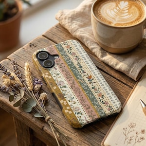 May include: A floral patterned phone case with vertical stripes in shades of cream, pink, green, and gold. The case is on a wooden surface with a cup of coffee and dried flowers.