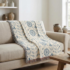 Puede incluir: Una manta crema con un estampado floral azul y flecos. La manta está sobre un sofá beige. El diseño floral incluye flores grandes y estilizadas y ramas con hojas. Los flecos son una mezcla de crema, azul y marrón.