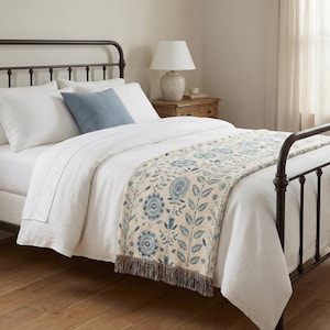 Pu&ograve; includere: Un letto con lenzuola bianche, un cuscino blu e un copriletto decorativo. Il copriletto presenta un motivo floreale blu e beige, con un bordo a frange. Una lampada &egrave; su un comodino di legno.