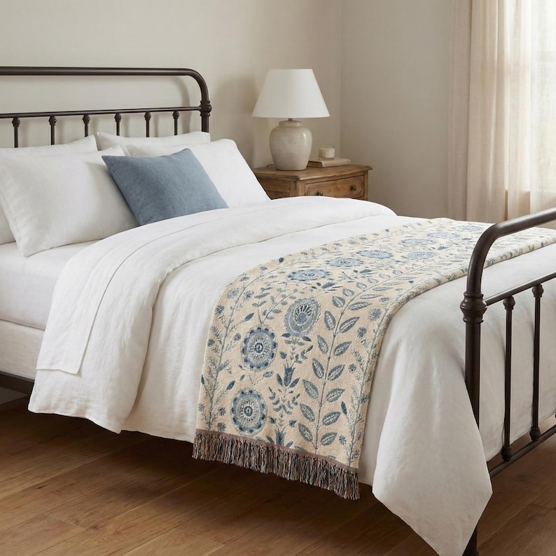 Pu&ograve; includere: Un letto con lenzuola bianche, un cuscino blu e un copriletto decorativo. Il copriletto presenta un motivo floreale blu e beige, con un bordo a frange. Una lampada &egrave; su un comodino di legno.