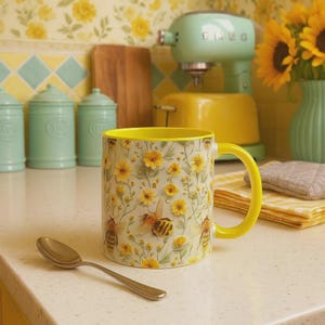 May include: A ceramic mug with a yellow handle and interior, featuring a floral and bee pattern. The mug is set on a white countertop with a spoon. The background includes a yellow and floral wallpaper, a mixer, and a vase with sunflowers.