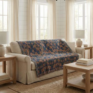 May include: A woven throw blanket featuring a dark blue background with a repeating pattern of orange and green floral motifs. The blanket has a fringed edge and is draped over a light beige sofa, displaying a textured fabric construction.