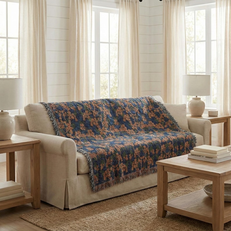 May include: A woven throw blanket featuring a dark blue background with a repeating pattern of orange and green floral motifs. The blanket has a fringed edge and is draped over a light beige sofa, displaying a textured fabric construction.
