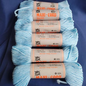 May include: Six spools of light blue braided Maxi-Cord yarn are stacked on a dark blue surface. Each spool has a label with the brand name, yardage, and other details. The yarn is labeled as brush-out and fade-resistant.