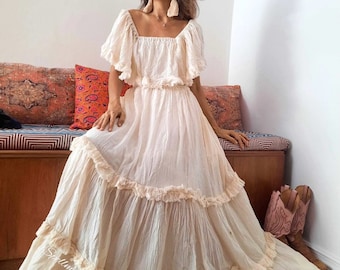 May include: A white maxi dress with a square neckline and short sleeves. The dress has a tiered skirt with lace trim and a ruffled bodice. The dress is made of a lightweight fabric and has a flowy silhouette.