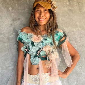 May include: A woman wearing a handmade off-the-shoulder top adorned with fabric flowers in shades of teal and pink. She is also wearing a tan crocheted hat with a flower accent, a decorative belt, and denim shorts. The outfit has a bohemian aesthetic.