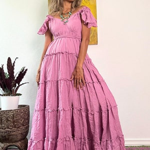 May include: A long, flowing, dusty rose-colored dress with off-the-shoulder sleeves and a tiered skirt. The dress features a fitted bodice and a full, ruffled skirt. A turquoise necklace adds a pop of color. The dress is suitable for a special occasion.