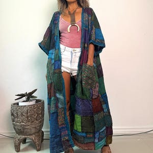 May include: A long, open-front patchwork duster coat featuring a variety of blue, green, and purple fabric squares with diverse patterns. The garment includes two large front pockets, three-quarter length sleeves, and a loose, flowing silhouette.
