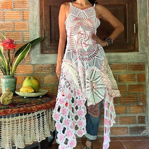 May include: A white and pink crochet dress with a unique design. The dress features a floral pattern and a flowing, asymmetrical hemline. The model is wearing blue jeans and green sandals.