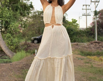 May include: A woman wearing a white, floor-length, halter-style dress with a cut-out design and a tie at the waist. The dress has a ruffled hem and is made of a lightweight fabric.