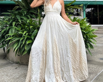May include: A white maxi dress with a lace trim. The dress has a halter neckline and a fitted bodice. The model is wearing the dress with a flower in her hair.