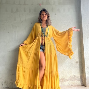 May include: A long, flowing mustard yellow robe with bell sleeves and a ruffled hem. The robe is open in the front and tied at the waist. The sleeves have a ruffled edge. The robe is made of a lightweight fabric.