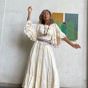 May include: A white cotton maxi skirt with a tiered design and a ruffled hem. The skirt is paired with a white cotton crop top with ruffled sleeves. The model is wearing a beaded necklace and a belt with a blue and white pattern.