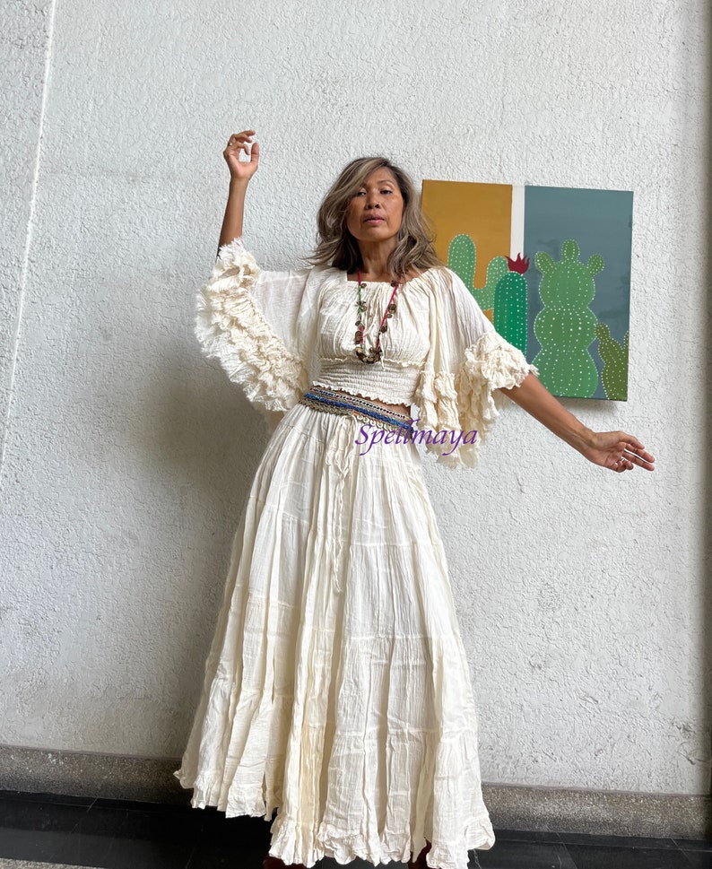 May include: A white cotton maxi skirt with a tiered design and a ruffled hem. The skirt is paired with a white cotton crop top with ruffled sleeves. The model is wearing a beaded necklace and a belt with a blue and white pattern.