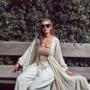 May include: A woman in a beige tube top, wide-leg trousers, and a cream-coloured kimono jacket, wearing black sunglasses. She is seated on a wooden bench, with a green hedge in the background. The outfit is a casual, bohemian style.