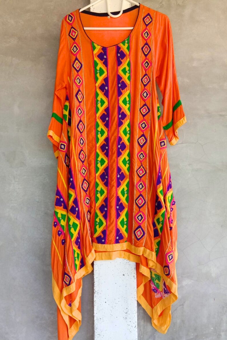 May include: A long, orange dress with colorful embroidery. The dress has a high neckline and long sleeves. The embroidery is in a geometric pattern and includes purple, green, yellow, and pink thread. The dress is asymmetrical and has a long, flowing hemline.